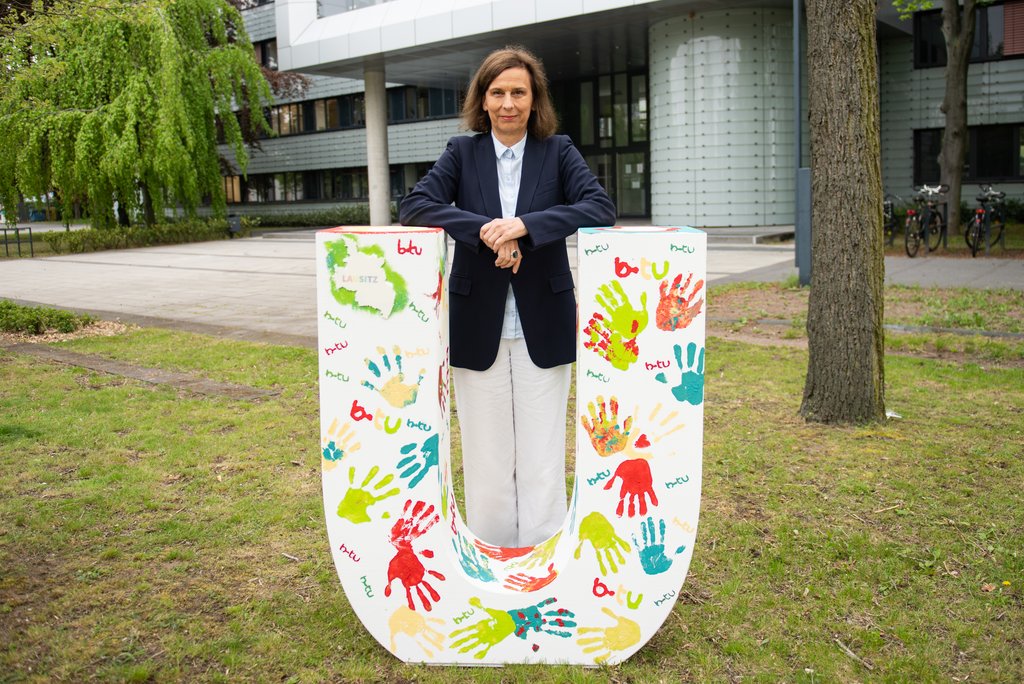 Die BTU-Präsidentin mit einem mit bunten Händen bemalten Buchstaben "U" vor dem Hauptgebäude der Universitätu