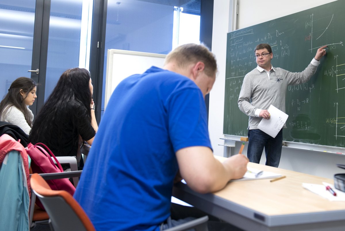 Künftige Studierende lösen, an Tischen in einem Seminarraum sitzend, Aufgaben in einem Mathematik-Vorkurs an der BTU Cottbus–Senftenberg. Der Dozent gibt, an der Tafel stehend, Erläuterungen.