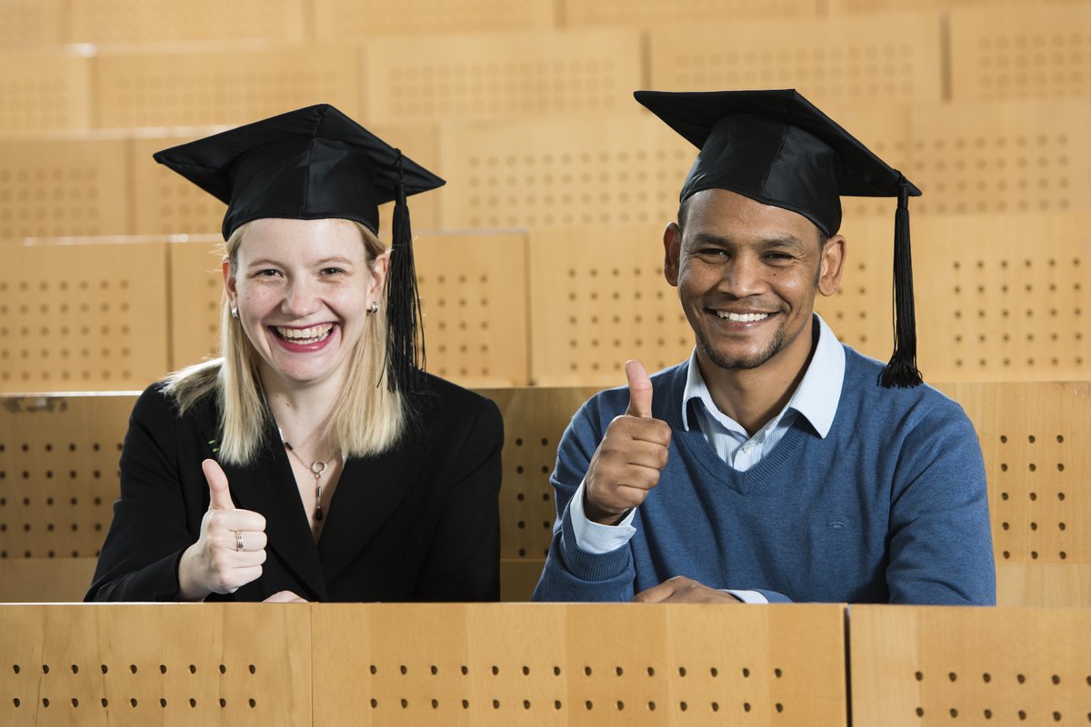 Zwei Studierende mit Doktorandenhüten sitzen lachend im Hörsaal und zeigen einen Daumen-hoch.