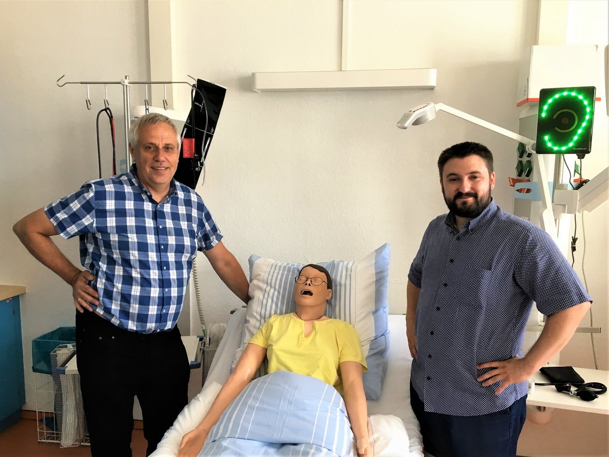 Der Studiengangsleiter des Studiegangs Pflegewissenschaft Florian Schimböck und Gastprofessor Dr. Thomas Boggat stehen im modernen Skills Lab der BTU rechts und links neben einer im Krankenbett liegenden Pflegepuppe.