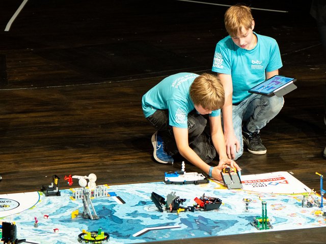 Zwei Schüler aus dem FIRST LEGO League-Team der BTU präsentieren ihr Projekt, hinter der Spielfläche kauernd. 