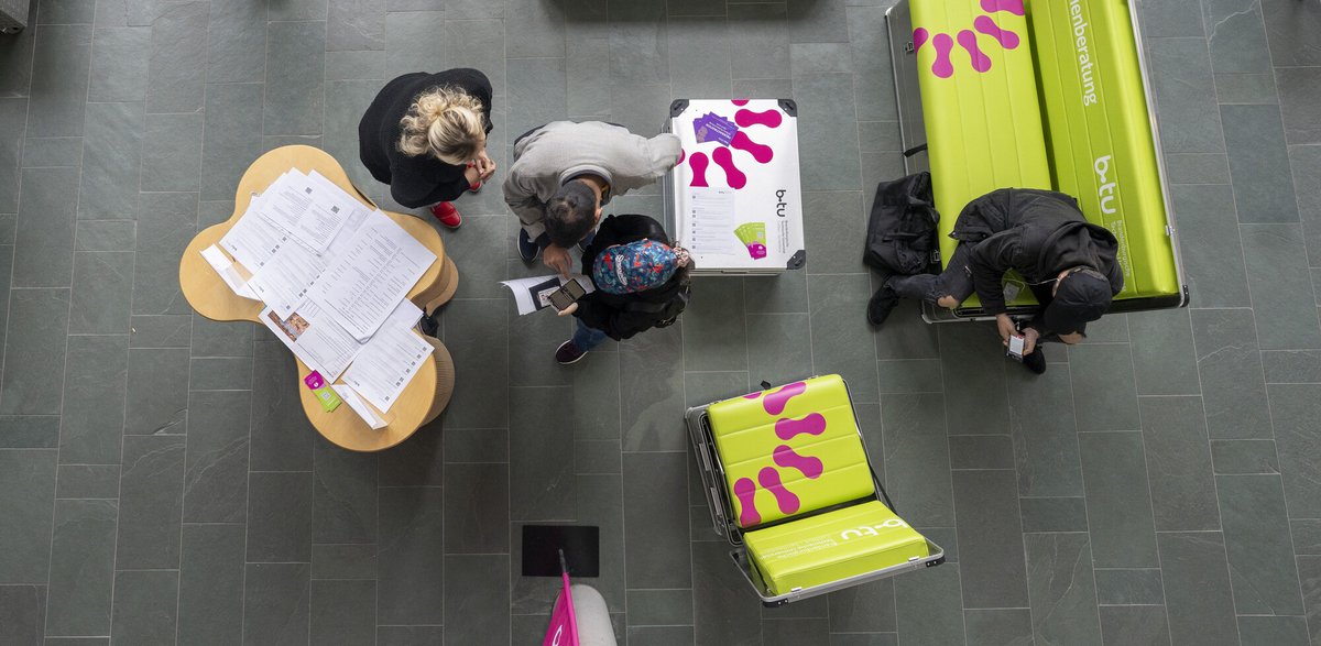 Das Foto zeigt den InfoPoint von oben. Zu sehen sind ein kleiner Tisch, zwei Sitzbänke und drei junge Menschen.