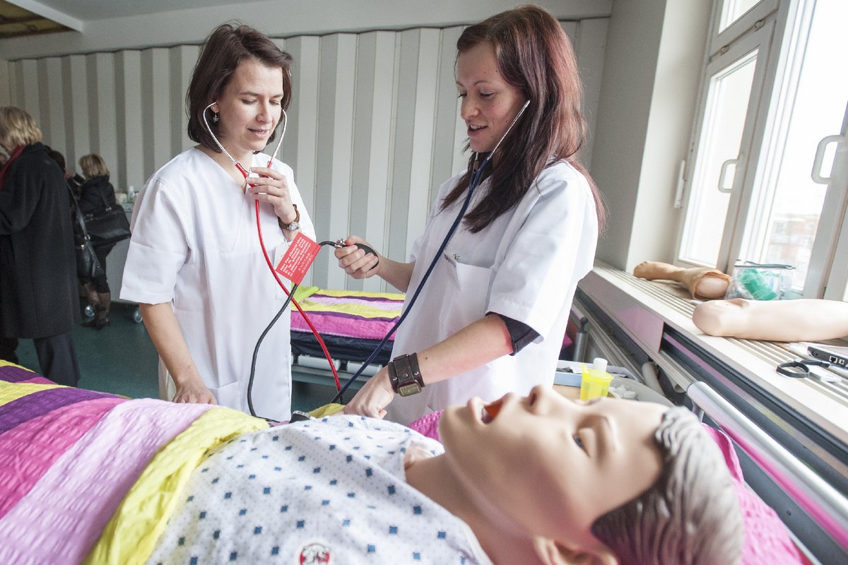 Zwei Studentinnen der Pflegewissenschaft stehen mit Behandlungsinstrumenten an einem Krankenbett, in dem eine Pflegepuppe liegt.