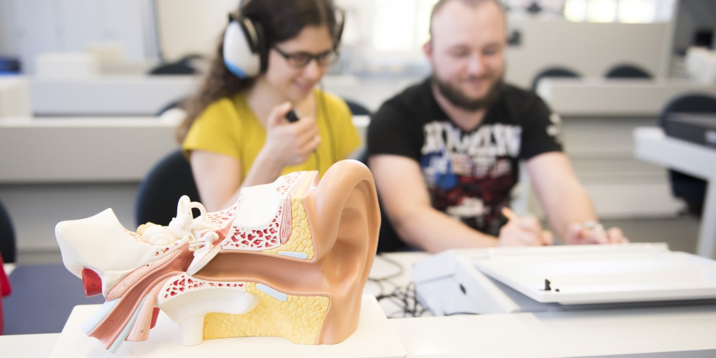 Im Vordergrund ein Modell des menschlichen Ohrs, im Hintergrund unscharf Studierende bei einem Hörtest