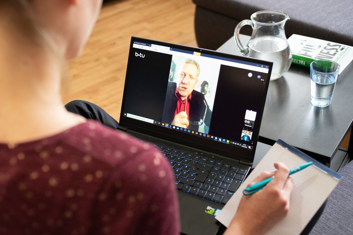 Eine weibliche Person, mit dem Rücken zum Betrachter, arbeitet im Rahmen einer Videokonferenz an einem Laptop. 