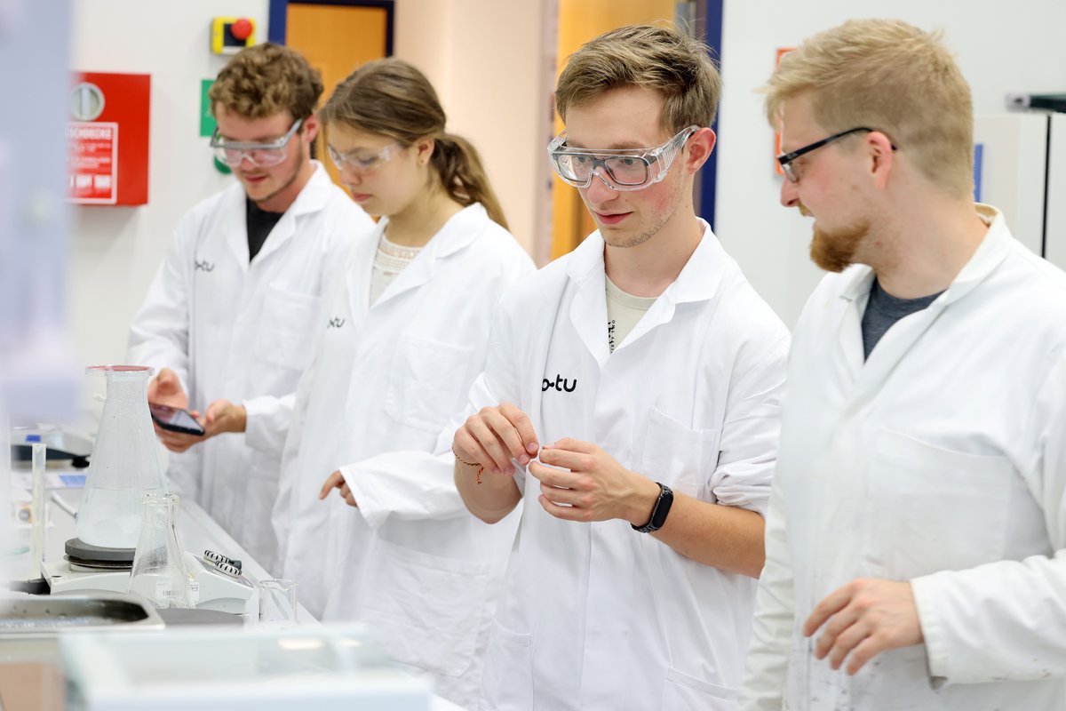 Eine Schülerin und drei Schüler stehen in weißen Kitteln in einem Chemielabor der BTU und experimentieren.