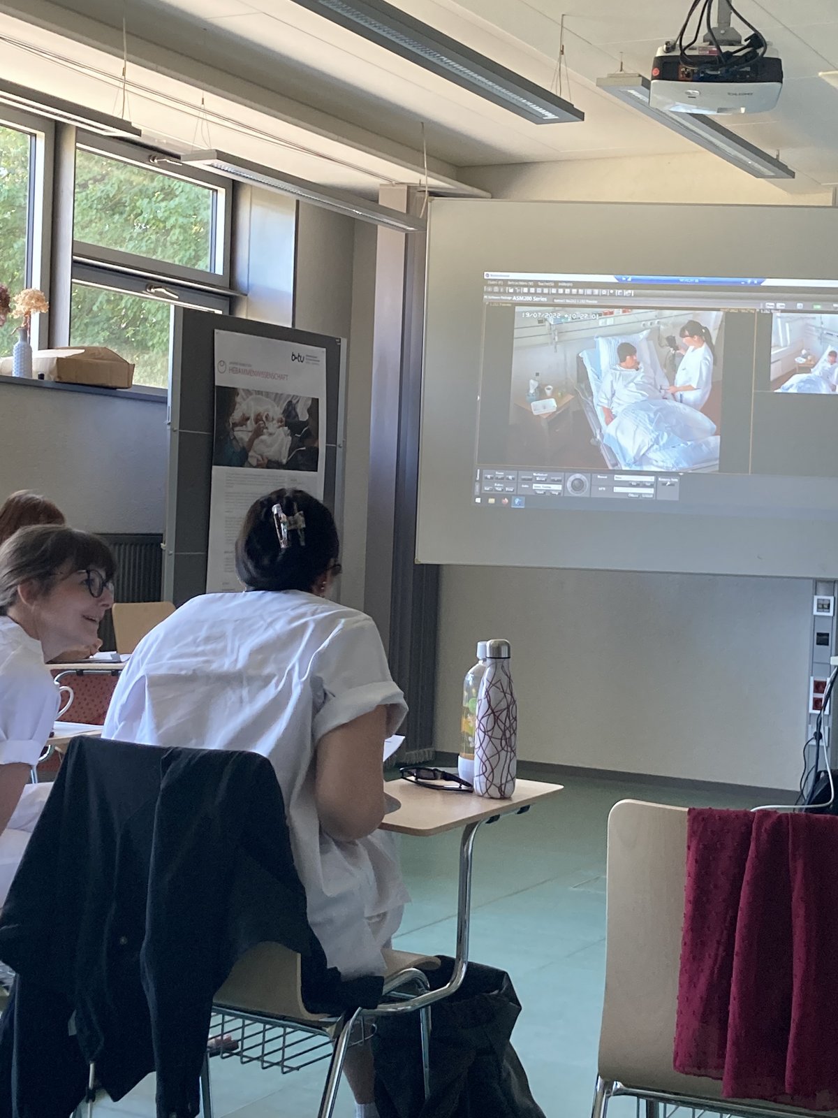 zwei Studierende beraten sich am Tisch und beobachen dabei die Simulationsübertragung per Videoleinwand