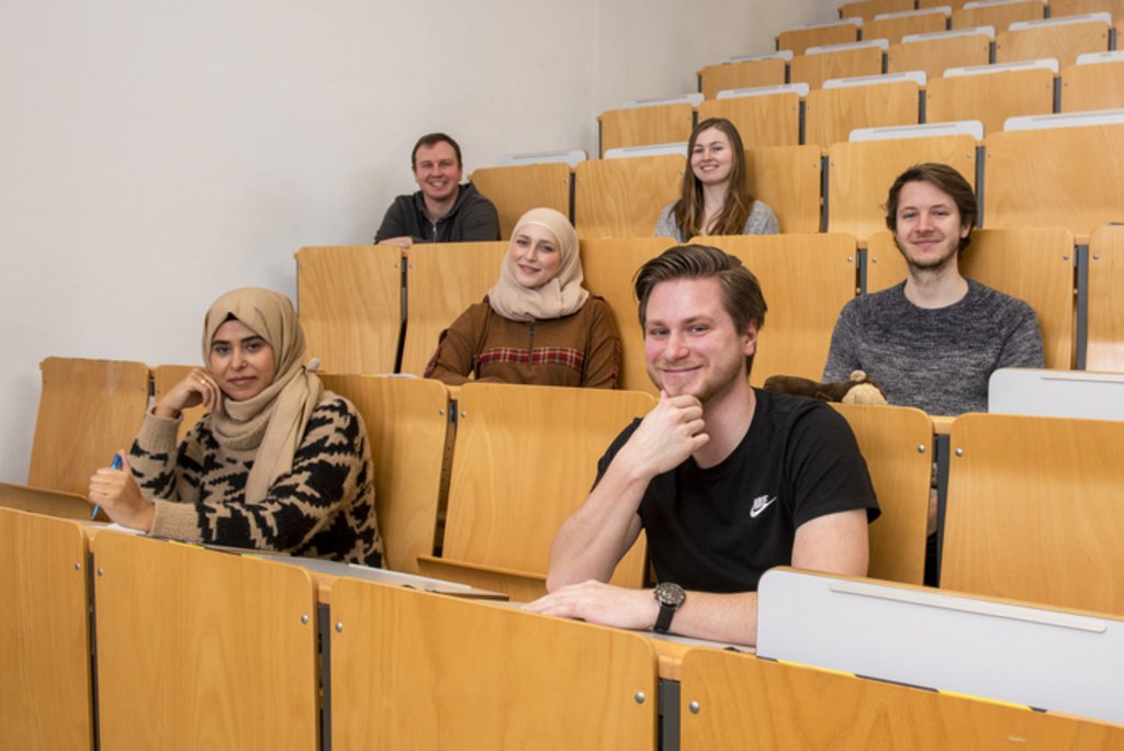 Mehrere weibliche und männliche Studierende unterschiedlicher Herkunft sitzen im Hörsaal und lächeln in die Kamera