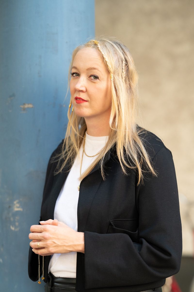 Porträt von Frau Prof. Verena Issel mit langen blonden Haaren und schwarzem Blazer vor einem zweifarbigen Hintergrund.
