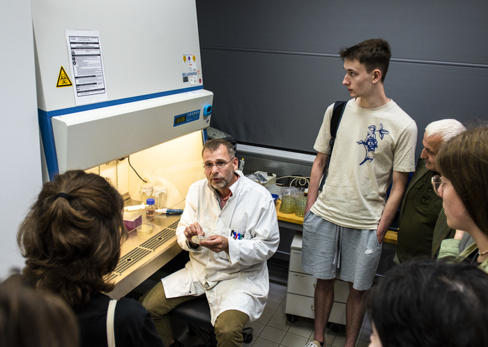 Eine Schülergruppe steht in einem Labor um einen sitzenden Dozenten im weißen Kittel