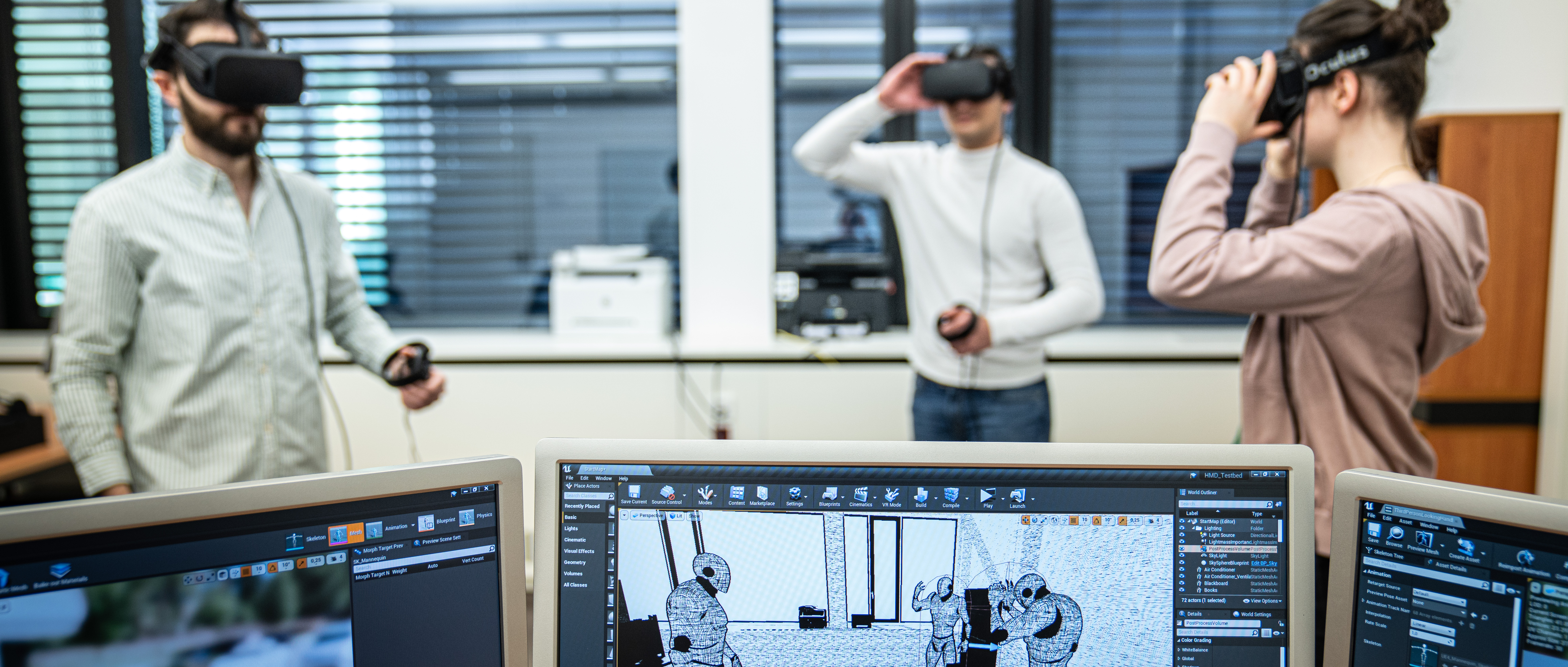 Drei Studierend, stehend im Raum, haben VR-Brillen auf, während auf einem PC-Monitor die passenden Avatare im virtuellen Raum zu sehen sind