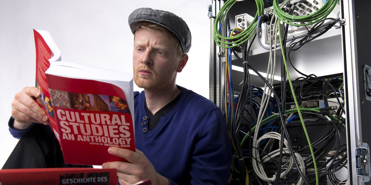 Student in the Master in Culture and Technology sits in front of a switchboard and reads a book with the title "Cultural Studies - an Anthology"