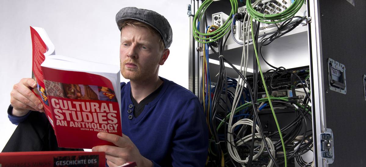 Werbebild für dem Master Kultur und Technik: Mann liest vor einem Kabelschrank ein Buch über Cultural Studies
