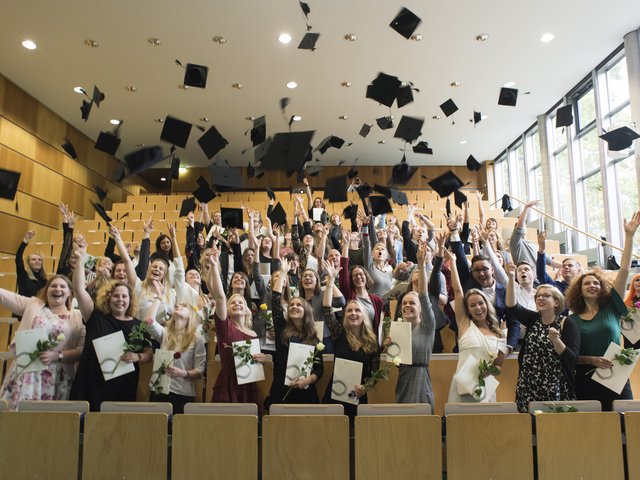 Absolventen und Absolventinnen im Hörsaal werfen ihre Alumni-Hüte in die Luft
