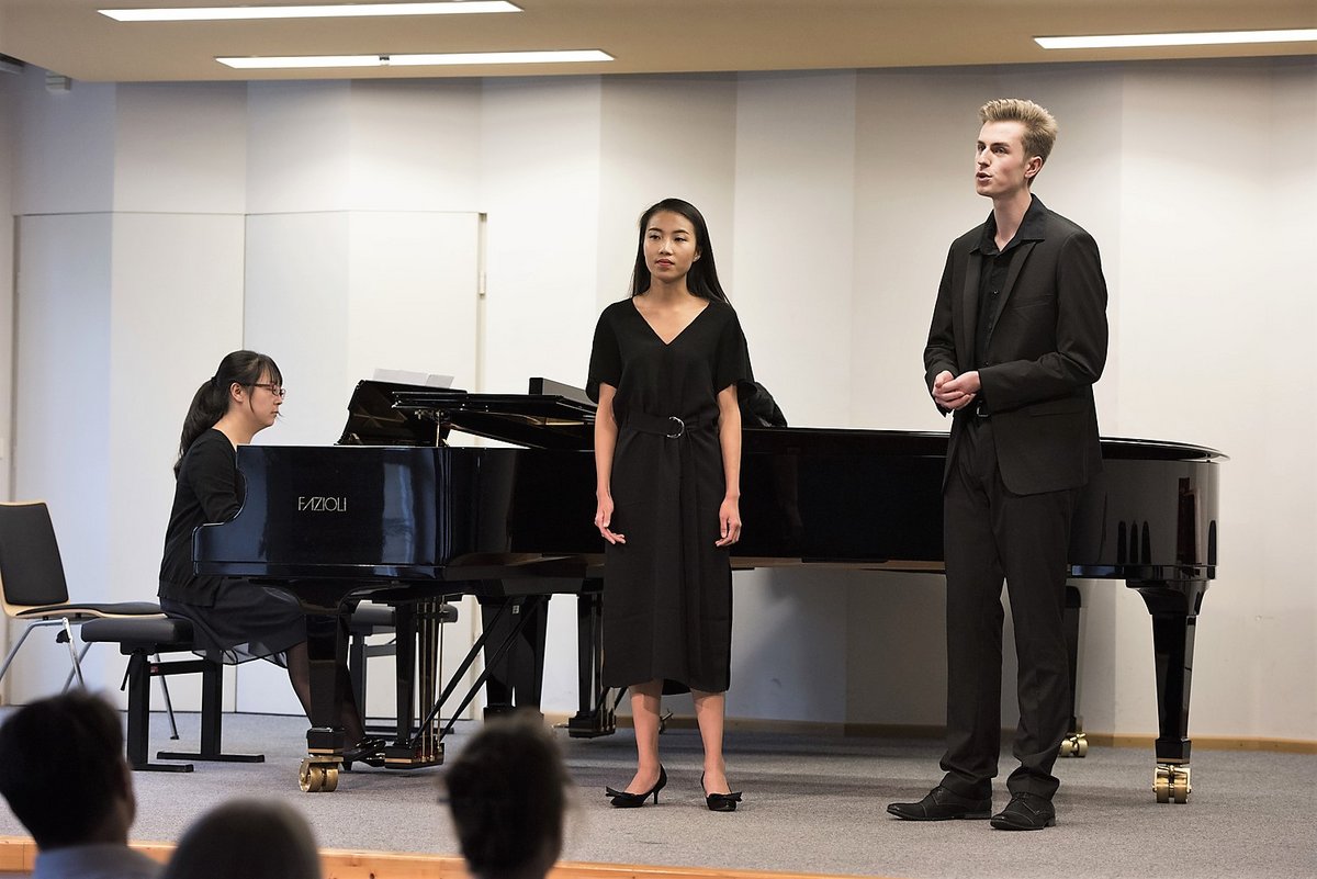 Auf eienr Bühne stehend, singen zwei Studierende, eien Student spielt sitzend Klavier.
