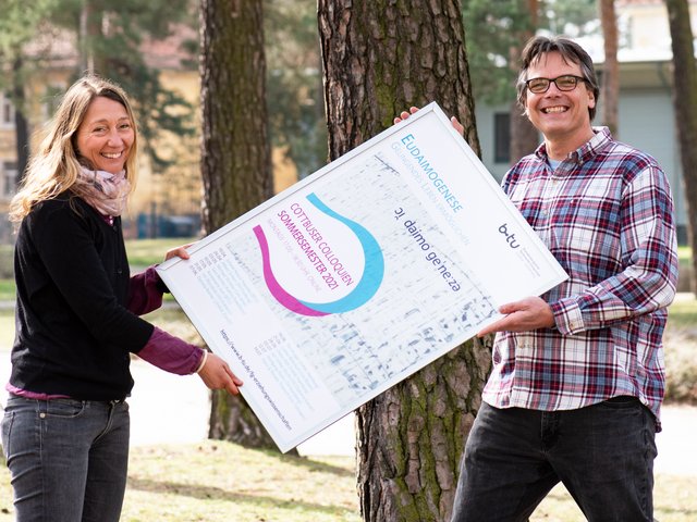 Juliane Noack Napoles und Thorsten Heimann halten ein Plakat der Veranstaltung Eudaimogenese - Gelingendes Leben ermöglichen zwischen sich