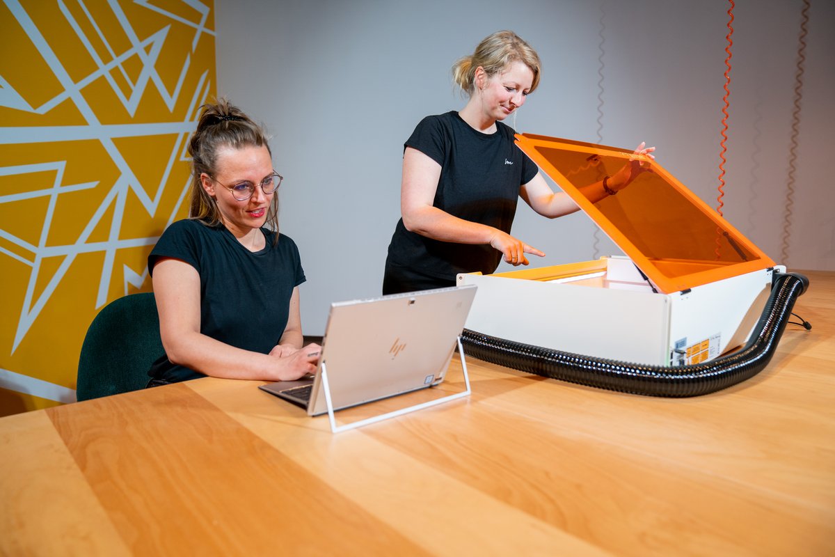 Dieses Foto stellt zwei Mitarbeiterinnen des COLab dar, von denen eine links am Laptop und eine rechts an weißem Lasercutter mit orangenen Lichtschirm der Marke Mr. Beam arbeitet.