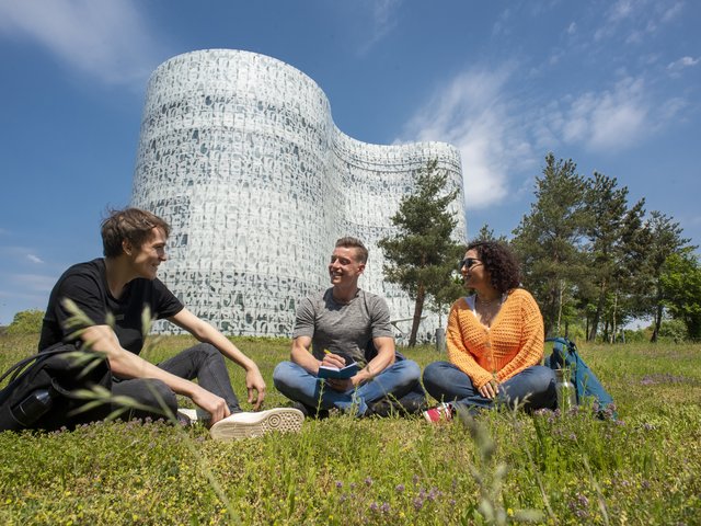 Three students sitting on a summer meadow in front of the Information, Communication and Media Centre at BTU.