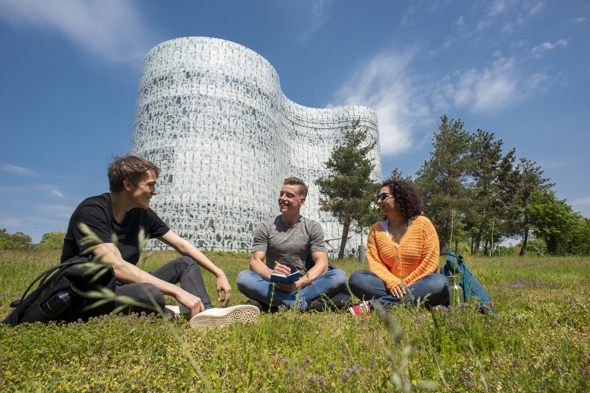 Three students sitting on a summer meadow in front of the Information, Communication and Media Centre at BTU.