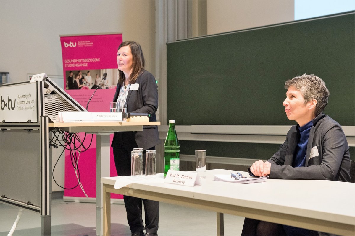 Prof. Dr. Anja Walter (li.) spricht, am Rednerpult stehend,  zu den Teinehmenden, daneben, am Tisch sitzend:  Prof. Dr. Heidrun Herzberg vom Institut für Gesundheit der BTU.