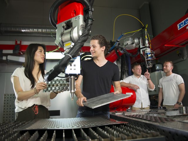 Studierende stehen links un rechts von eienm Roboterarm und untersuchen Werkstücke aus Metall.