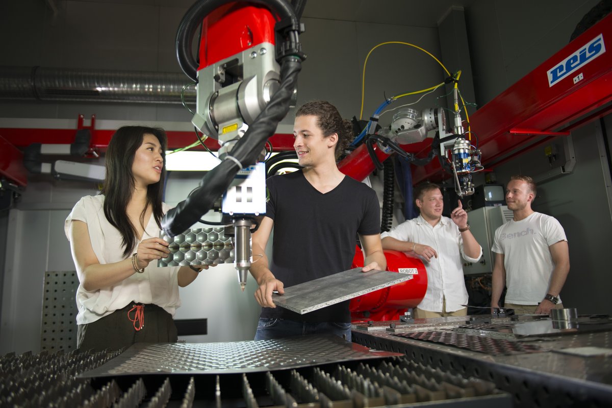 Studierende stehen links un rechts von eienm Roboterarm und untersuchen Werkstücke aus Metall.