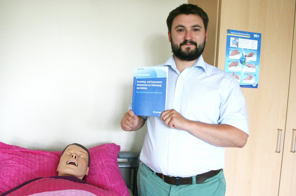 Florian Schimböck mit seiner Buchveröfentlichung im Skills Lab des Studiengangs Pflegewissenschaft am Pflegebett einer Simulationspuppe.