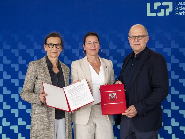 Drei personen (zwei Frauen links und ein Mann rechts) stehen vor einer blau-grünen Messewand. Die Frauen halten ein aufgeschlagenes Buch in der Hand. Der Mann ein geschlossenes rotes Buch..