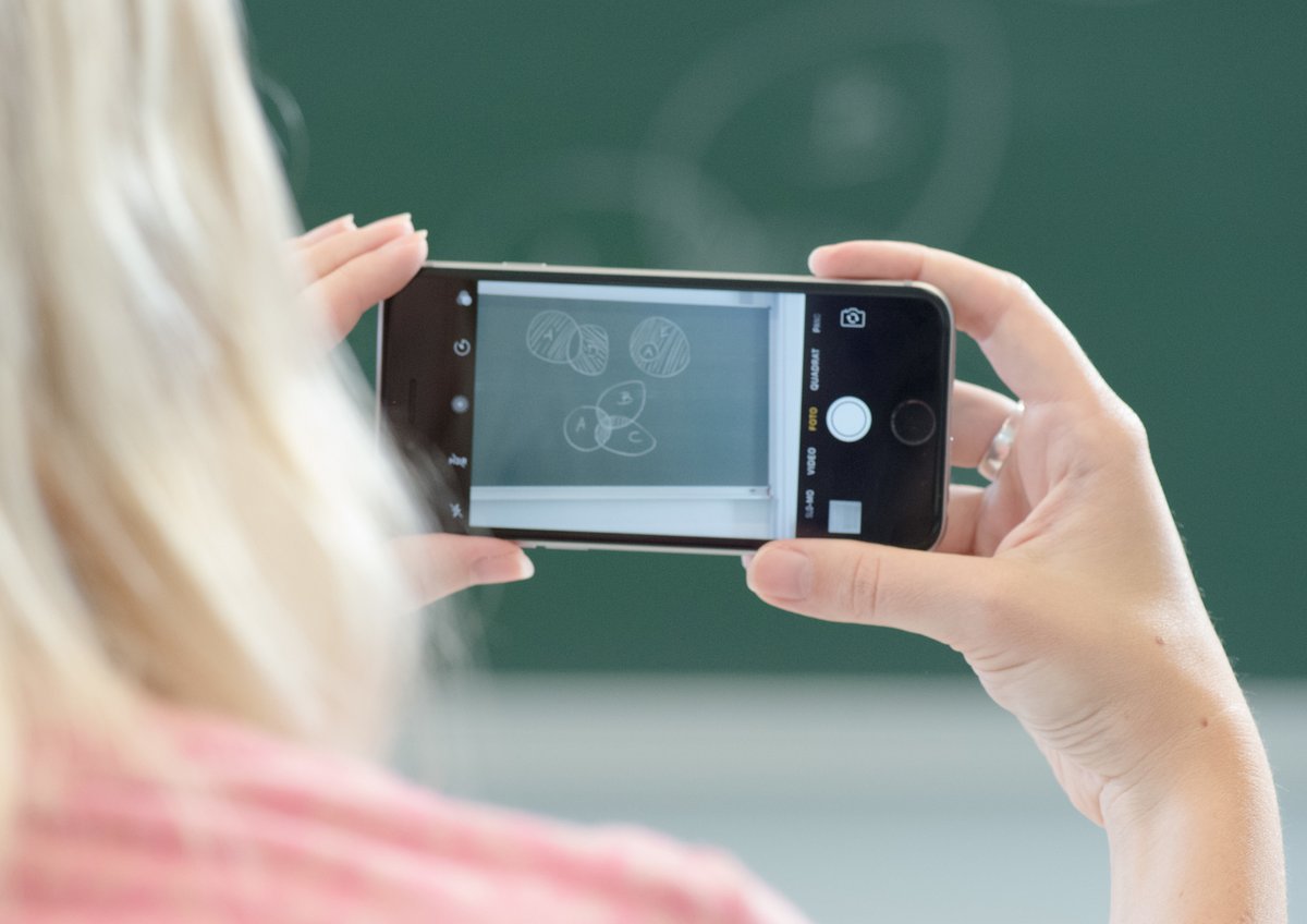 Mit dem Smartphone hält eine weibliche Person, mit dem Rücken zum Betrachter stehend, auf einer Tafel abgebildete Lehrinhalte fest. 