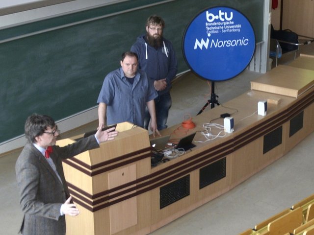Der wissenschaftliche Mitarbeiter Stephan Hernschier (Li.) und zwei weitere Personen geben, im Hörsaal sthend, Erläuterungen zur akkustischen Kamera.