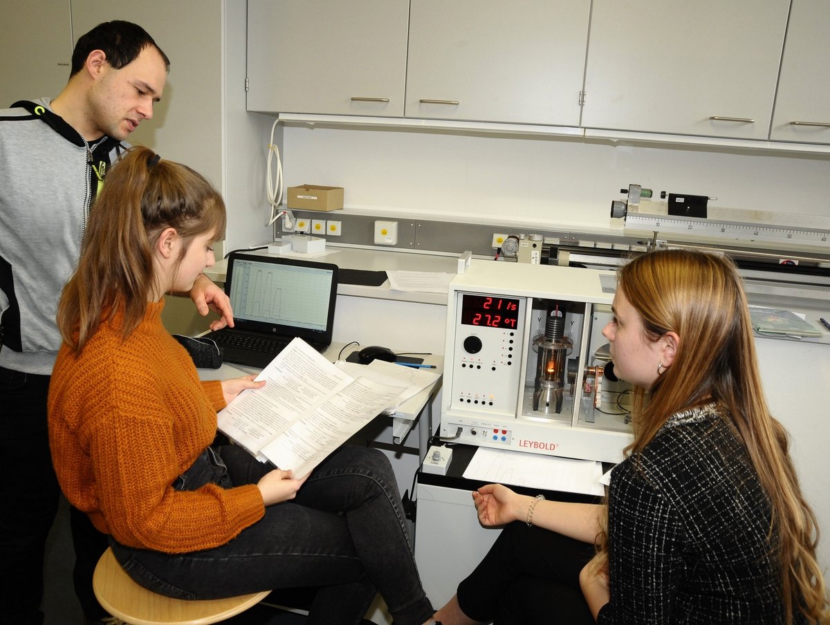 Laboringenieur Shadi (inks) spricht stehend mit den vor einem Schulröntgengerät sitzenden Abiturientinnen Amy Fritz und Luzie Richter (v. li.).