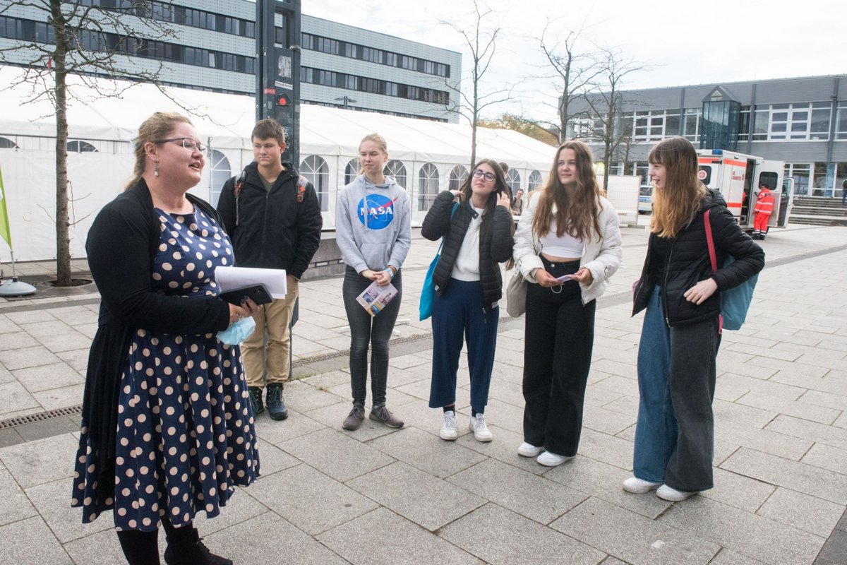 Mädchen und Jungen stehen auf dem Campus und folgen den Ausführungen einer Mitarbeiterin der Universität.