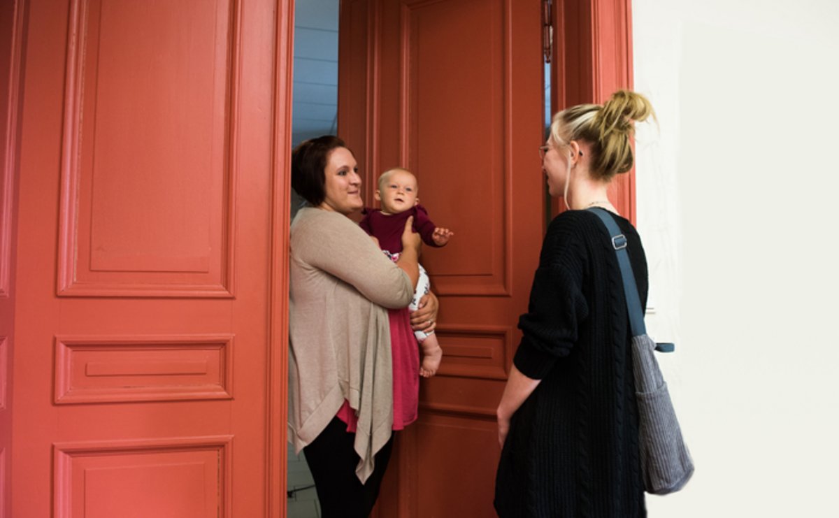 Das Foto zeigt eine Frau mit Baby auf dem Arm, die an der Haustür eine Sozialarbeiterin empfängt.