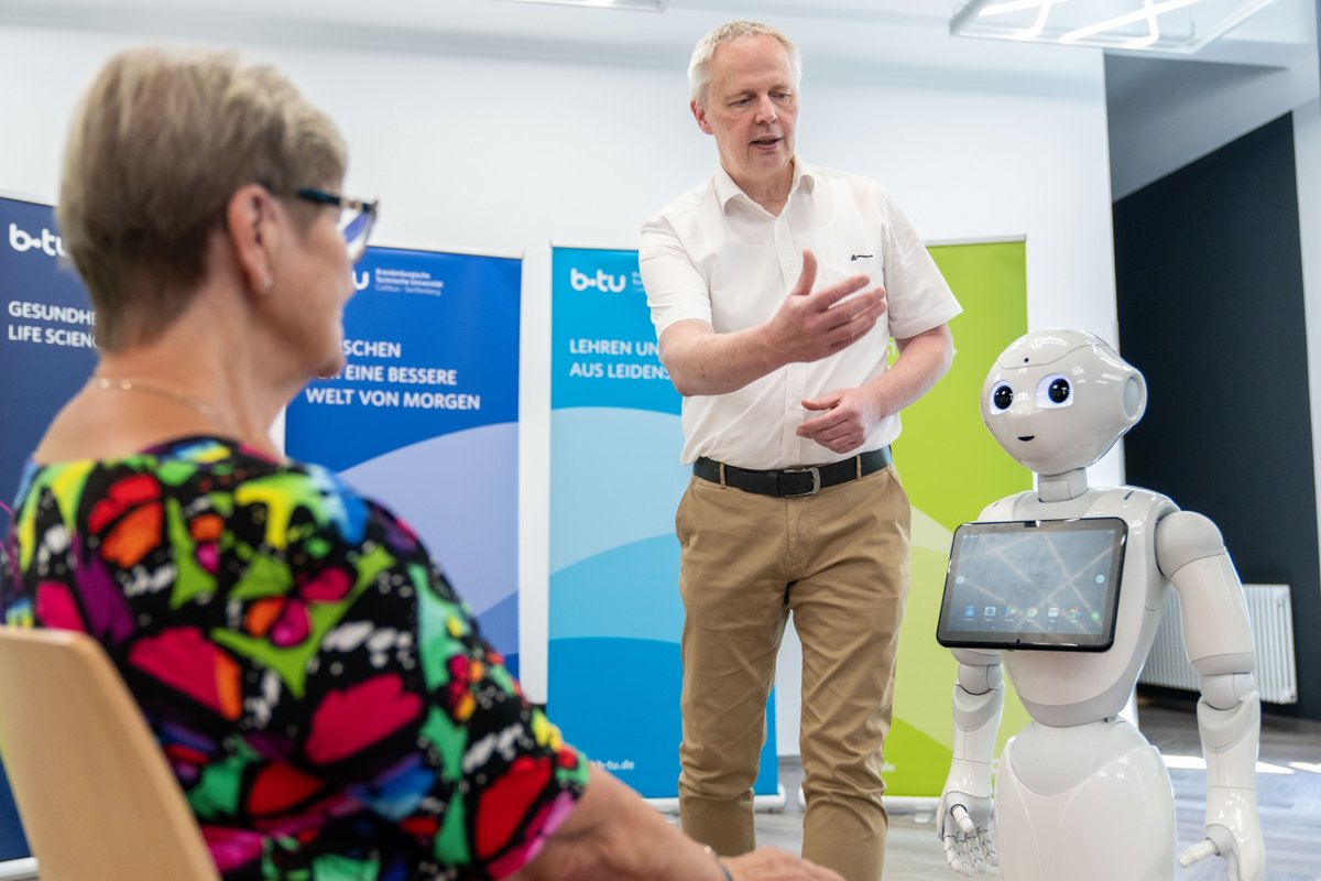  Simulation patient sitting in dialog with the robot Pepper. BTU nursing scientist Jörg Matthäi gives explanations while standing. 