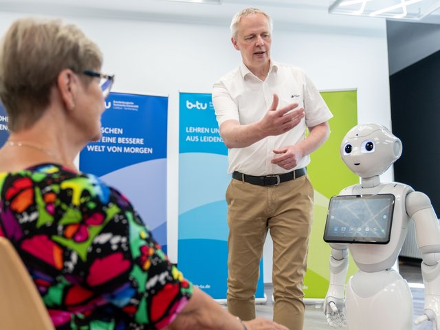  Simulationspatientin sitzend im Dialog mit dem Roboter Pepper. Der BTU-Pflegewissenschaftler Jörg Matthäi gibt stehend Erläuterungen. 