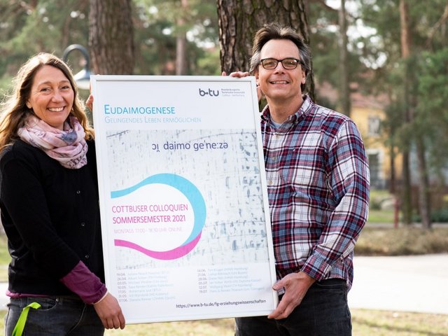 Prof. Juliane Noack Napoles und der wissenschaftliche Mitarbeiter Thorsten Heimann halten das Veranstaltungsplakat