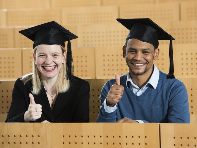 Zwei Studierende mit Doktorhüten im Hörsaal sitzend.