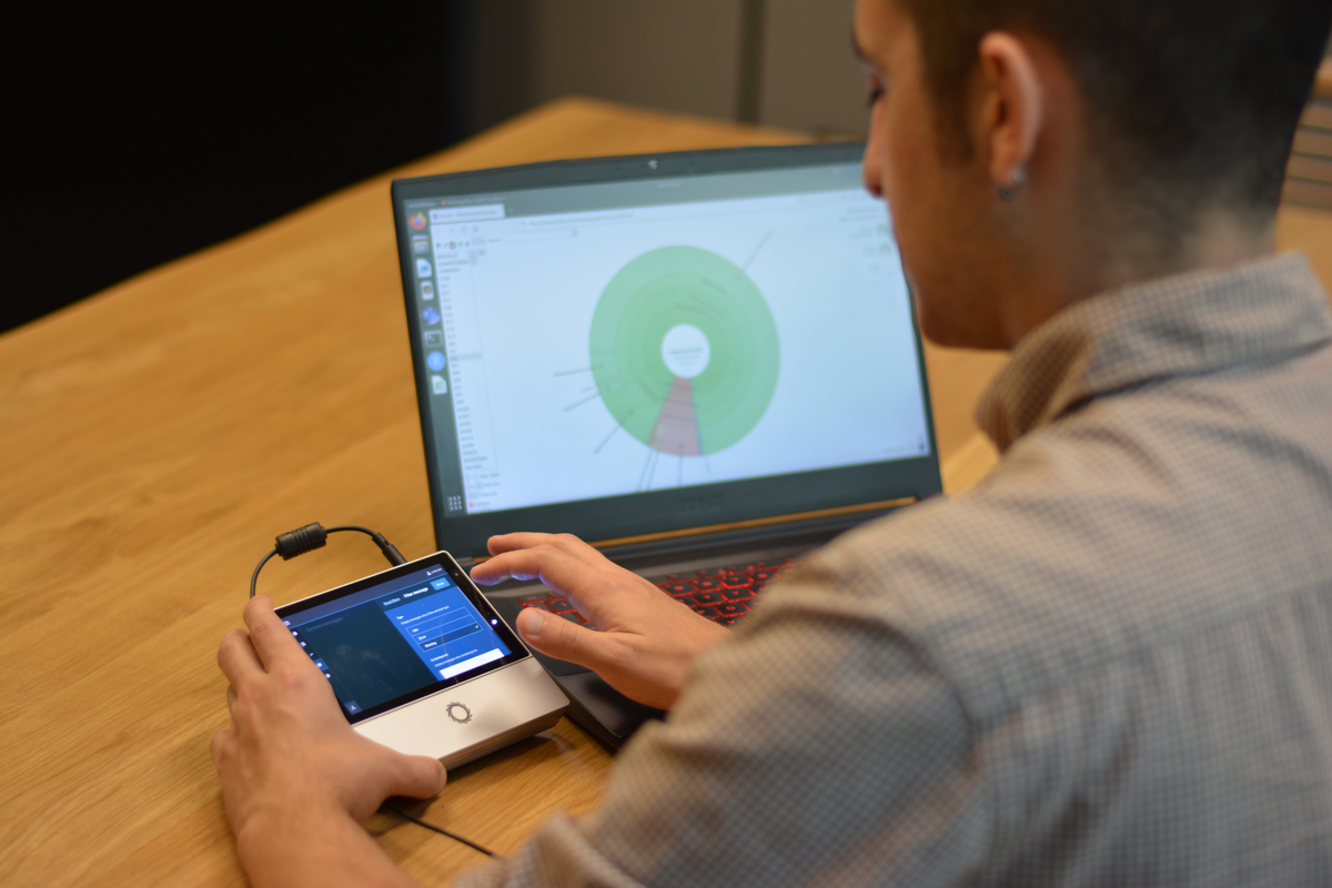 Adriel Latorre (Direktor der Genomikabteilung bei Darwin Bioprospecting) im Umgang mit einem DNA-Sequenzierer (Modell MinION). Im Hintergrund die Visualisierung eines Krona-Plots auf einem Laptop, die das taxonomische Profil einer der in der Studie entnommenen Schlammproben zeigt