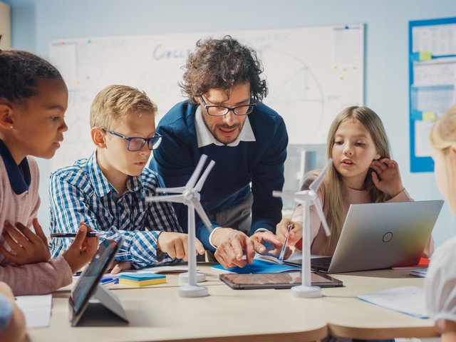 Das Foto zeigt eine Lehrkraft an einem Tisch mit vier Schüler*innen bei einer Gruppenarbeit mit Laptop.