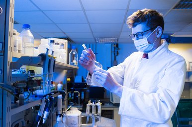 Wissenschaftler im weißen Laborkittel und mit Schutzmaske arbeitet im Labor an einer Probe zwischen Laborgeräten.