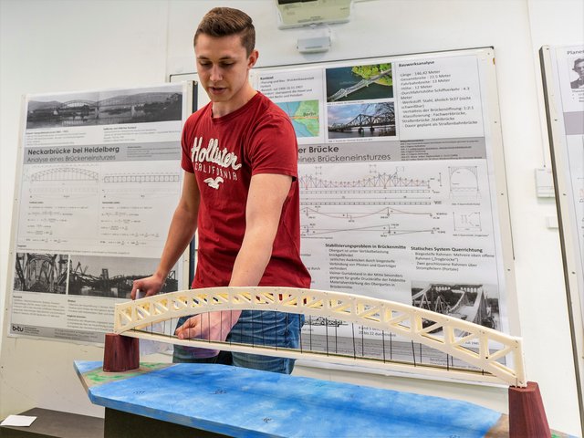 Ein Student gibt, hinter dem Modell einer Brücke stehend, Erläuterungen zu den Ursachen realer Brückeneinstürze.