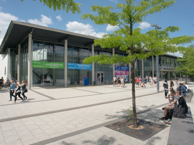 Studierende stehen vor dem Zentralen Hörsaalgebäude, andere sitzen auf Bänken vor dem Campus.