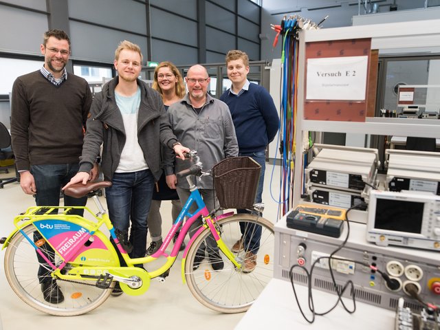 Personengruppe steht im Elektrotechnischen Labor und blickt in die Kamera. Robert Ebel präsentiert das bunte BTU-Fahrrad