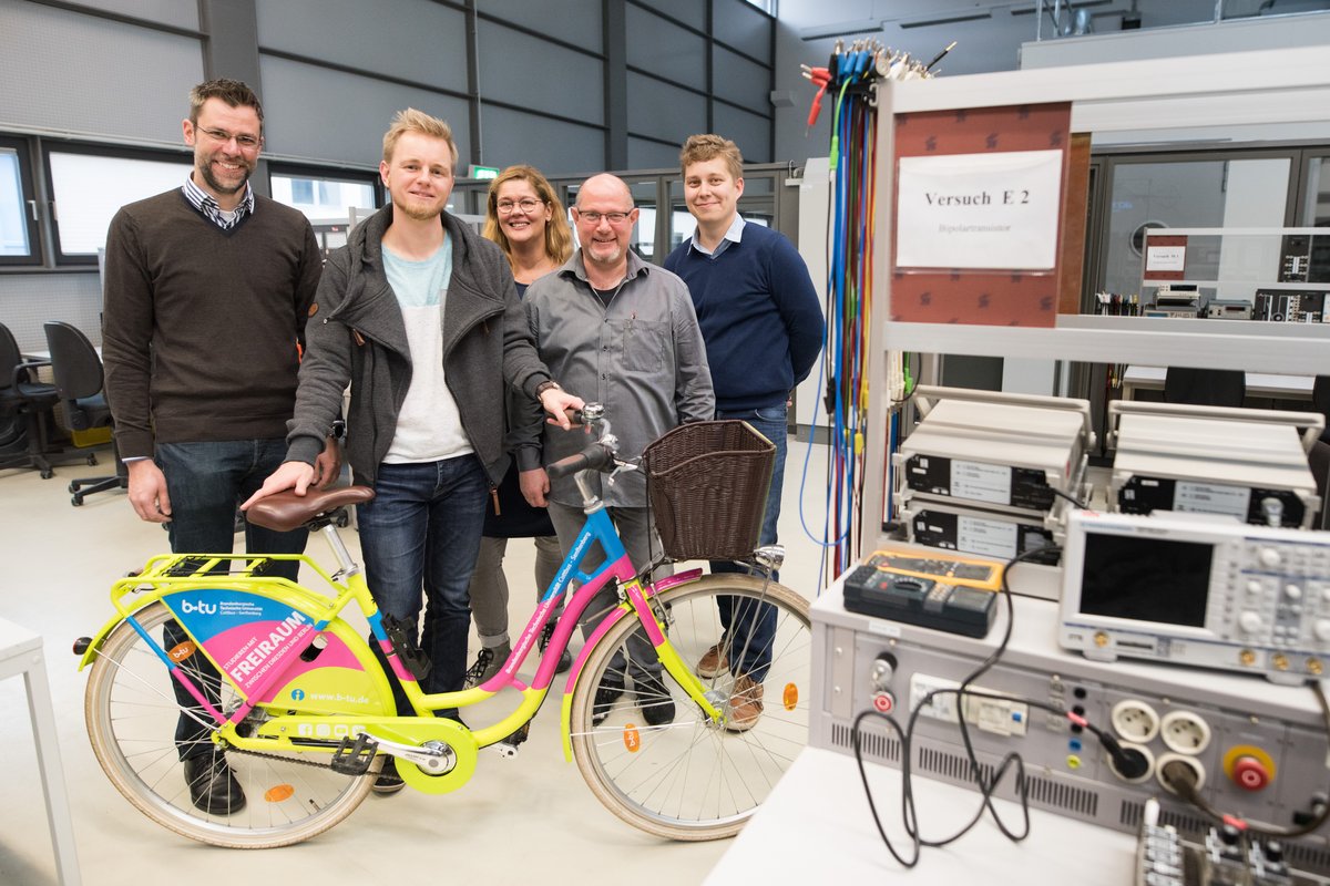 Personengruppe steht im Elektrotechnischen Labor und blickt in die Kamera. Robert Ebel präsentiert das bunte BTU-Fahrrad