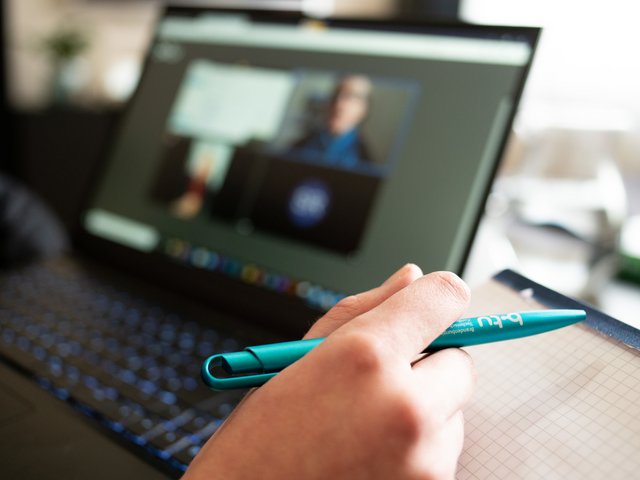 Das Foto zeigt eine Hand einer am Laptop sitzenden Person in einer Videokonferenz.mit einem Kugelschreiber.