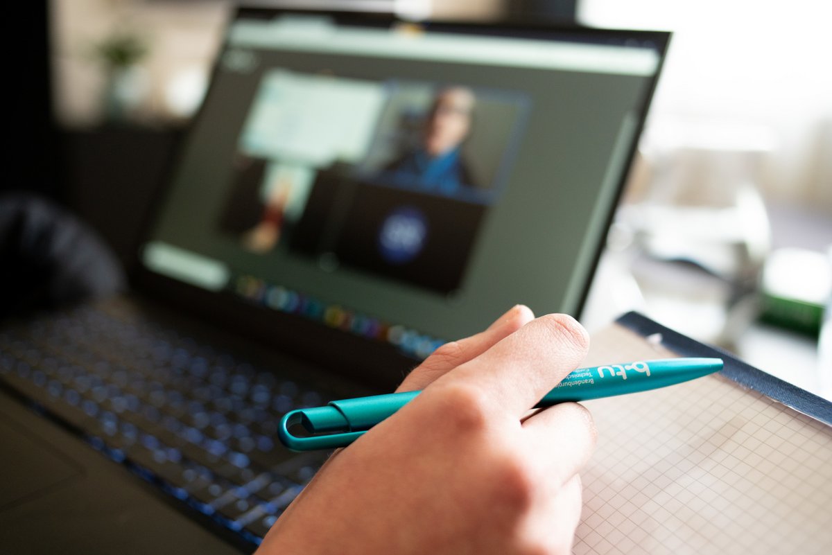 Das Foto zeigt eine Hand einer am Laptop sitzenden Person in einer Videokonferenz.mit einem Kugelschreiber.