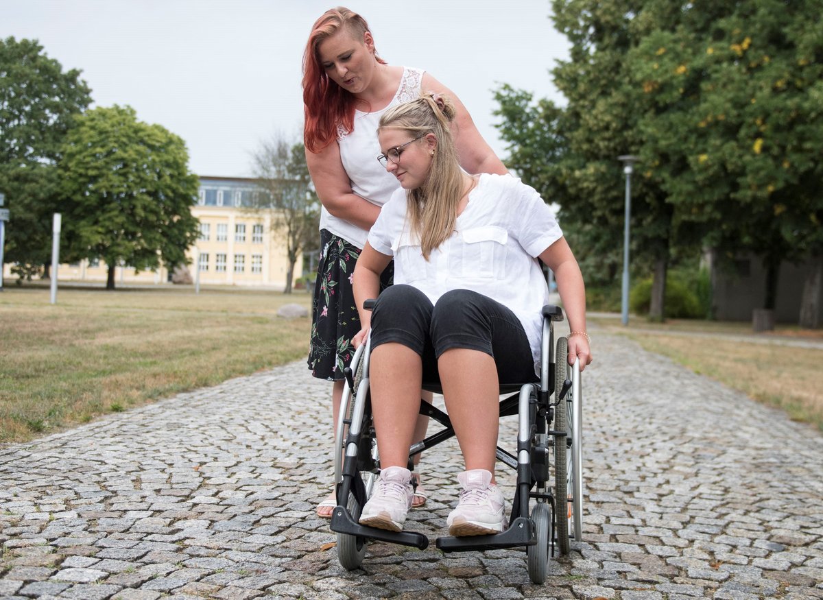 Zwei Studentinnen, eine im Rollstuhl sitzend, eine hinter ihr stehend, prüfen auf Kopfteinpflaster die Möglichkeit, sich auf diesem fortzubewegen. prüfen die Möglichkeit, sich per Rollstuhl auf Kopfsteinpflaster fortzubewegen.