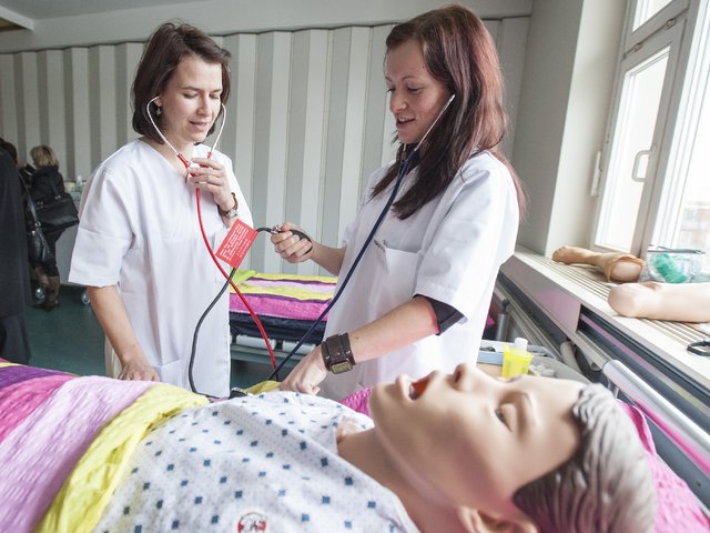 Zwei Studentinnen der Pflegewissenschaft stehen mit Behandlungsinstrumenten an einem Krankenbett, in dem eine Pflegepuppe liegt.