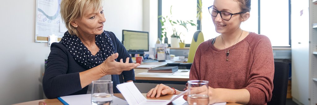 Beratung 2 Frauen sitzen zusammen in einem Büro an einem Tisch, auf dem Broschüren liegen. Die linke Frau spricht zur rechten Frau..