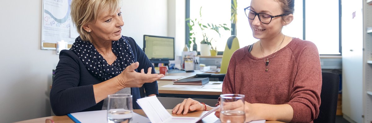 2 Frauen sitzen zusammen  in einem Büro an einem Tisch, auf dem Broschüren liegen. Die linke Frau spricht zur rechten Frau.. 